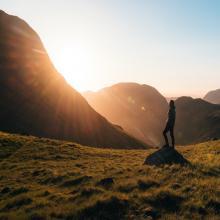 Silhouette photography of person standing on green grass in front of mountains during golden hour